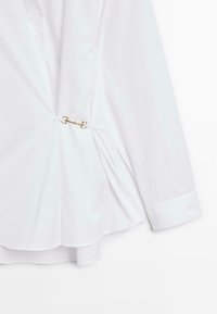 Chemise blanche à manches longues avec un détail décoratif de fermoir en mors de cheval doré à la taille, dotée d'un ourlet évasé et de poignets boutonnés.