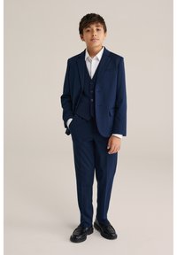 Mörkblå kostym i tre delar med enkelknäppt kavaj, matchande byxor och vit skjorta. Svarta skor fullbordar outfiten.