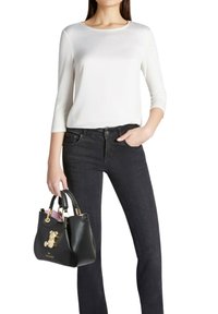 Borsa a tracolla in pelle nera con accessori dorati, impreziosita da un ciondolo a forma di orsetto. Indossata con una camicia bianca a maniche lunghe e jeans neri.