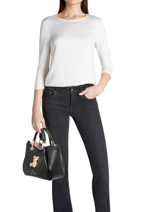 Borsa a tracolla in pelle nera con accessori dorati, impreziosita da un ciondolo a forma di orsetto. Indossata con una camicia bianca a maniche lunghe e jeans neri.