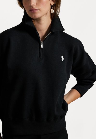 Svart quarter-zip hettegenser laget av myk stoff, med hevet krage, ribbestrikkede ermer, og en liten hvit brodert logo på brystet.