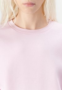 Felpa rosa chiaro con scollatura rotonda e tessuto morbido e texture, mostrata dalla vita in su. Design semplice e monocolore.
