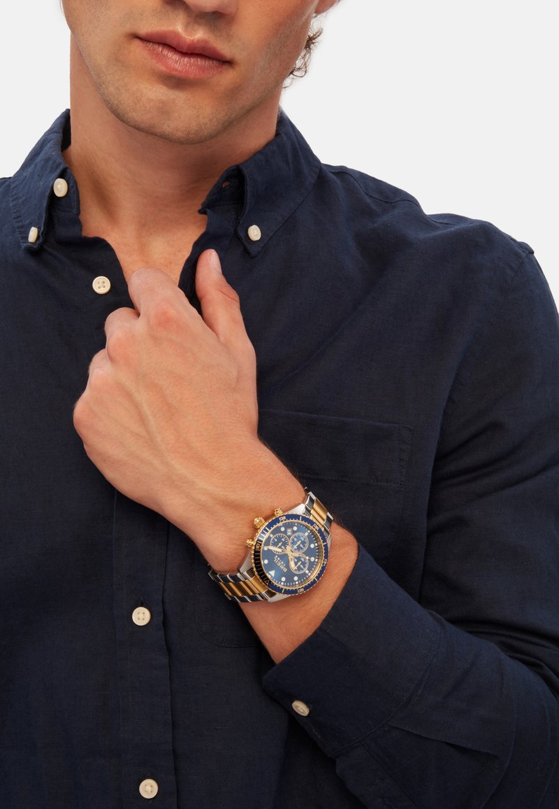 Montre chronographe bleue avec des accents dorés, bracelet en acier inoxydable et cadran bleu texturé. Portée au poignet, accompagnée d'une chemise noire à boutons.