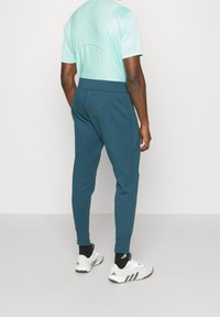 Man med teal joggers, ljusblå kortärmad skjorta, svarta strumpor och vita Adidas-sneakers, stående mot en enkel vit bakgrund.