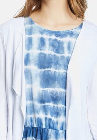 Weißer offener Cardigan über einem blauen Batik-Top. Das Top weist unterschiedliche Blautöne auf, hat eine weiche Textur und einen rundlichen Halsauschnitt.
