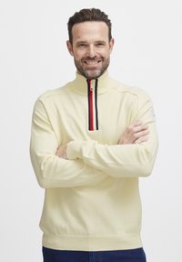 Fény sárga pulóver széles gallérral, rövid cipz�árral és piros-fekete-fehér csíkkal a elülső részen. Pamut keverék anyag.
