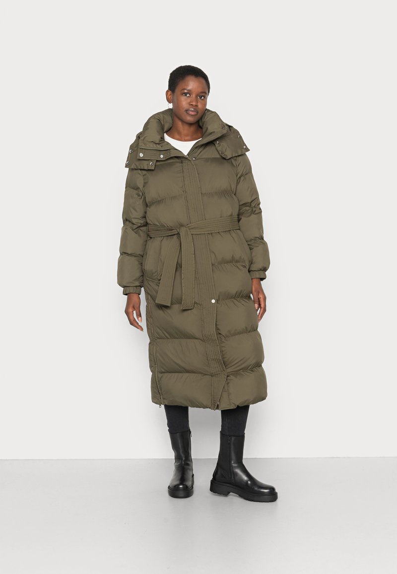 Casaco oversized acolchoado em verde oliva, com cintura cingida, gola alta e capuz destacável. Textura suave e bolsos laterais. Botas pretas.