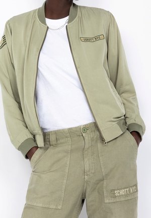 Persoon draagt een licht olijfgroene Schott NYC-jas en bijpassende broek met handen in de zakken over een wit T-shirt.