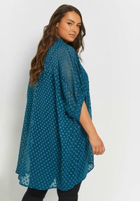 Kimono leggero color teal con motivo a pois, vestibilità ampia e maniche corte. Il tessuto ha una struttura trasparente e cade morbido.