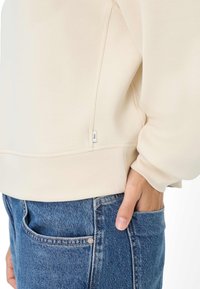 Crème kleur sweatshirt met een ronde halslijn, ribbelboorden en een label aan de zijnaad pocket. Gecombineerd met lichtblauwe denim jeans.