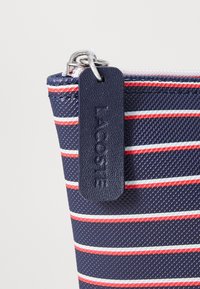 Tengerészes tasak vörös és fehér csíkokkal, textúrált felülettel, ezüst cipzárral, és bőr címkével, amelyen az "Lacoste" felirat van.