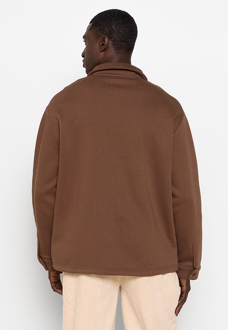 Homme portant une veste marron à manches longues et un pantalon beige clair, tourné le dos à la caméra devant un fond blanc uni.