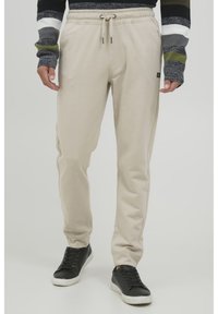Beige sweatpants med snor i taljen, tætsiddende ben og sidelommer, parret med sorte sneakers. Stoffet ser blødt og glat ud.