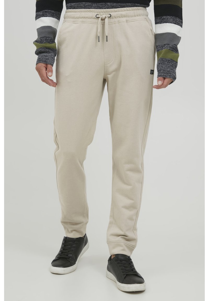 Beige sweatpants med snor i taljen, tætsiddende ben og sidelommer, parret med sorte sneakers. Stoffet ser blødt og glat ud.