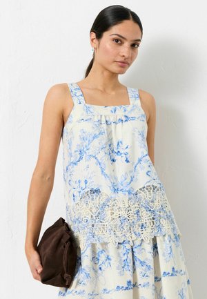 Frau trägt ein ärmelloses weiß-blau gemustertes Oberteil mit Blumenmuster und Spitzenbesatz, hält eine kleine braune Wildledertasche vor einer weißen Wand.