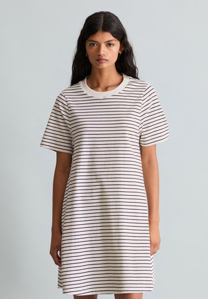 Vrouw met lang donker haar die een witte korte mouwen jurk draagt met dunne horizontale bruine strepen, staand tegen een effen lichtgrijze achtergrond.