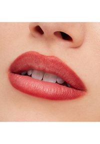 Zbliżenie ust pomalowanych matową różową szminką. Tekstura wygląda na gładką, z subtelnym połyskiem. Widoczne zęby, skóra o jasnym odcieniu.