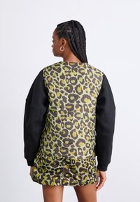 Løveprikket genser med grønn og brun design, med svarte ermer. Teksturert stoff med quiltet bakside.