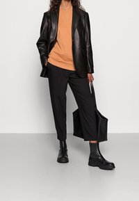 Persoon draagt een zwart leren blazer, oranje sweatshirt, zwarte cropped broek, zwarte laarzen en houdt een grote zwarte leren tote bag vast.