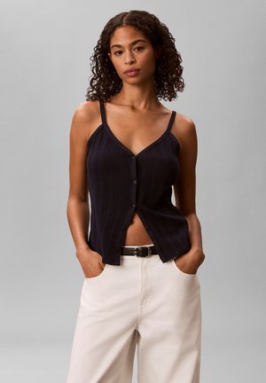 Młoda kobieta z kręconymi włosami, ubrana w czarny, zapinany top bez rękawów i kremowe spodnie, ręce w kieszeniach na szarym tle.