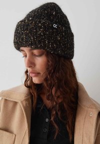 Beanie a maglia nera e marrone con un motivo testurizzato, con una piccola etichetta logo. Indossato con un cappotto beige e una maglietta nera sotto.