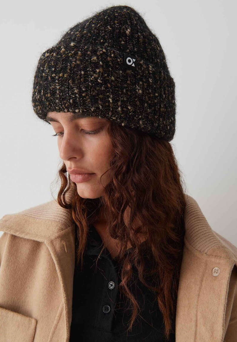 Beanie a maglia nera e marrone con un motivo testurizzato, con una piccola etichetta logo. Indossato con un cappotto beige e una maglietta nera sotto.