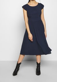 Nainen, jolla on yllään laivansinisestä midi-mekosta, jossa on liehuvat hihat, yhdistettynä mustiin nilkkakowboy-saappaisiin, seisoo jalat eristyksissä.