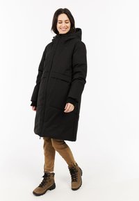 Abrigo acolchado negro con capucha, cremallera frontal, dos grandes bolsillos frontales y puños de canalé, combinado con pantalones beige y botas de senderismo marrones.