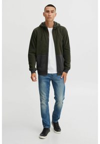 Sudadera con cremallera de felpa verde oliva con un panel gris en el dobladillo y bolsillos delanteros, combinada con jeans azul claro y zapatillas negras.