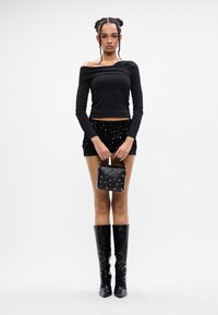 Joven con cabello trenzado que lleva una blusa negra sin hombros, una minifalda con lentejuelas y botas negras hasta la rodilla, sosteniendo un bolso negro con tachuelas.