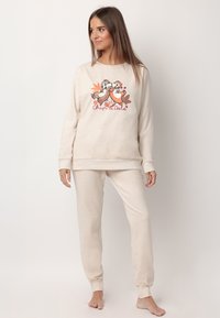 Mujer vestida con un conjunto de ropa para estar en casa de color beige, con un dibujo de Chip y Dale y hojas de otoño en el sudadera, de pie descalza frente a un fondo blanco.