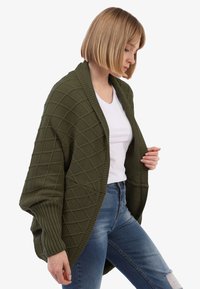 Olijfgroene gebreide cardigan met ruitpatroon, open voorkant, geribde manchetten en een ontspannen pasvorm, gedragen over een wit T-shirt en jeans.
