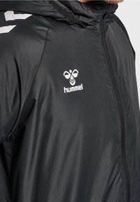 Närbild av en person som bär en svart zip-up jacka med huva och en vit hummel-logotyp på bröstet.
