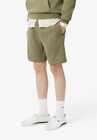 Pantalones cortos de algodón verde oliva con bolsillos laterales, combinados con una camisa de rayas claras y zapatillas blancas, que presentan detalles verdes y calcetines de tobillo acanalados.