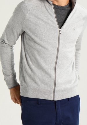 Mann trägt hellgrauen Reißverschluss-Cardigan über einem dunkelgrauen Hemd und marineblaue Hose, Hände seitlich an einem einfarbigen Hintergrund.