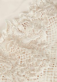 Tela de encaje en color beige claro con intrincados patrones florales y bordes en forma de scallop, que presenta una textura suave y un tejido detallado.