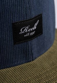 Dark blue corduroy cap with a ribbed texture, featuring a black label with "Reel" and "est. 1997." Brim is olive green with a similar texture.
