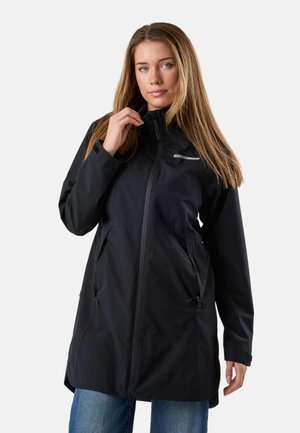 Woman wearing a long black waterproof jacket with zipped pockets and blue jeans, adjusting the jacket's collar.