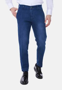 Pantaloni in denim blu con taglio dritto, tasche frontali e chiusura con bottone. Indossati con scarpe nere lucide e una camicia bianca.