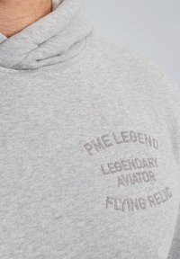 Sivi dukserica s teksturiranim završetkom. Sadrži izvezene natpise: "PME LEGEND", "LEGENDARY AVIATOR" i "FLYING RELIC" u tamnosivoj boji.