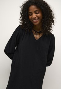Robe noire en tissu texturé avec un décolleté en V, des manches longues et un détail de nœud à la nuque. Motif froissé subtil sur l'ensemble.