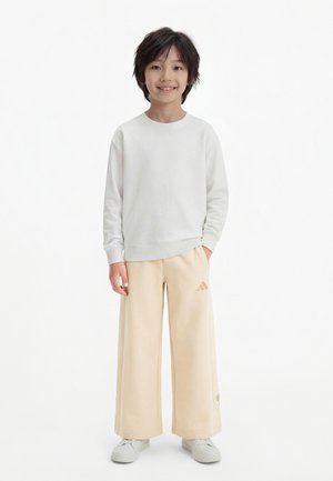 Šypsosi stovintis vaikas, vilkintis baltą ilgomis rankovėmis marškinėlį, smėlio spalvos plataus silueto kelnes su Adidas logotipu ir baltus sportinius batelius ant balto fono.