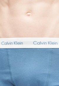 Ανδρικός κορμός που φορά μπλε εσώρουχο Calvin Klein, με εμφανές το λογότυπο στη ζώνη και εμφανές τον ομφαλό στο κέντρο της κοιλιάς.