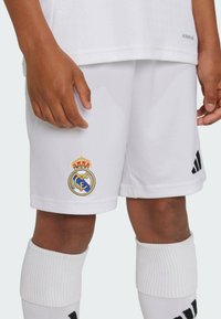 White soccer shorts with a gold-crowned Real Madrid logo, featuring an Adidas black accent on the side and textured fabric.