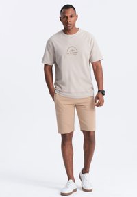 T-shirt in cotone beige con collo rotondo e maniche corte, con stampa a logo. Abbinato a pantaloncini beige e sneakers bianche.