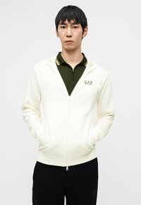 Κρέμ φούτερ με φερμουάρ μπροστά, φέρει λογότυπο στο στήθος και συνδυάζεται με πουκάμισο σκουροπράσινου χρώματος με γιακά από κάτω.