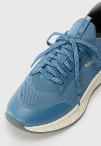 Blue athletic shoe with mesh and smooth synthetic material. Features round toe, textured accents, flat laces, and branded details on the side.