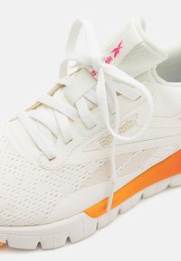 Biele športové topánky so sieťovaným zvrškom, šnúrkami a oranžovými akcentmi na podrážke. Obsahujú logo "Reebok" na boku a vzorovanú textúru.
