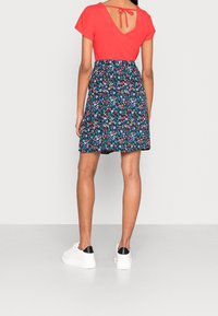 Robe fleurie avec un haut rouge et une jupe marine, ornée de fleurs rouges et bleues. L'arrière présente une découpe en V avec un détail noué. Stylisée avec des baskets blanches.