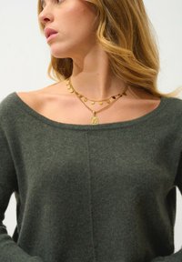 Dunkelgrüner, weicher Strickpullover mit einem schulterfreien Design. Begleitet von geschichteten goldenen Halsketten mit floralem und hängendem Akzenten.